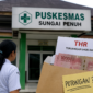 Seorang tenaga kesehatan berdiri di depan Puskesmas Sungai Penuh dengan ilustrasi amplop THR, menggambarkan dugaan potongan tunjangan dan tekanan terhadap pegawai.